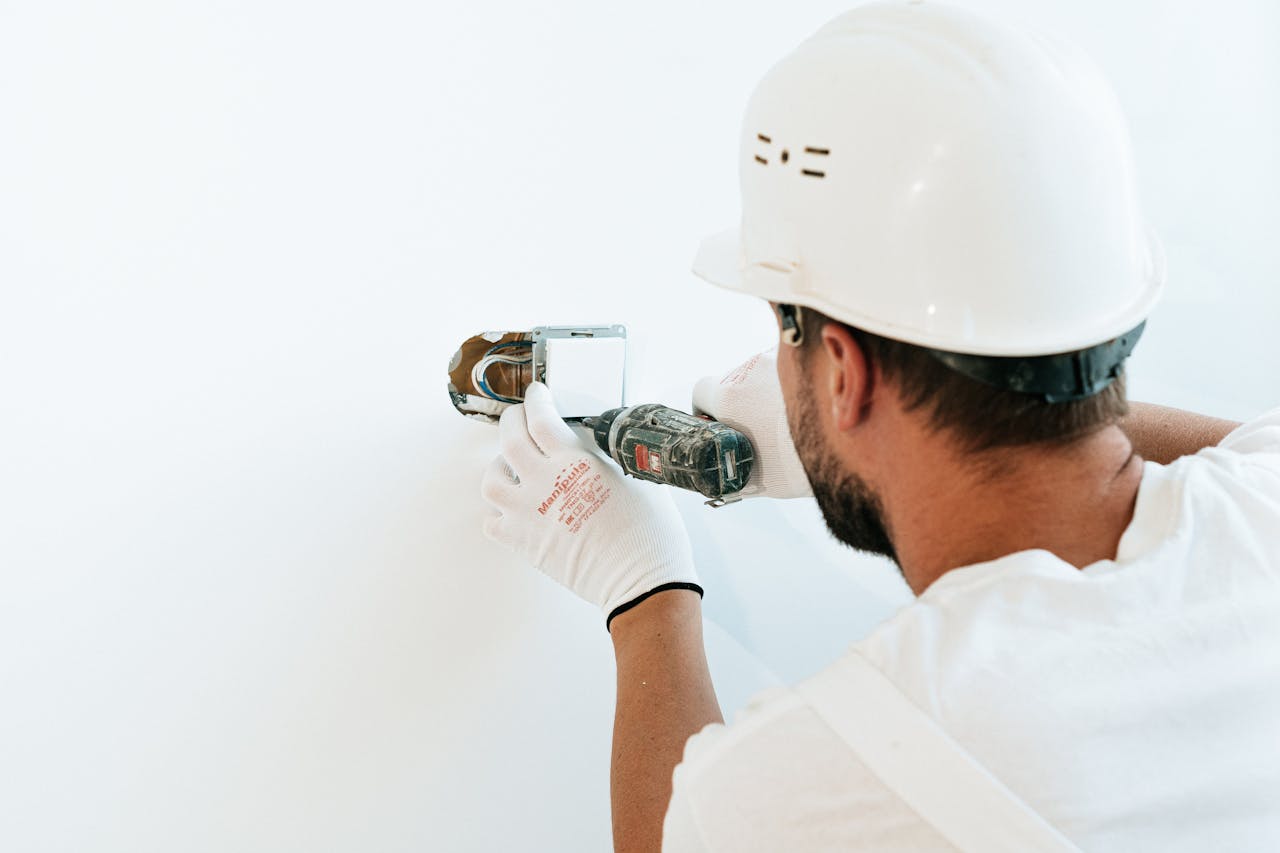 service-01 Construction worker wearing helmet and gloves uses a power tool to fix a wall.